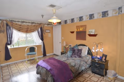 A bedroom with a single bed covered in patterned bedding and a purple throw blanket. There is a nightstand with a lamp and framed photo next to the bed. A window with sheer white curtains and dark drapes is on the left wall, with a chair placed in front of it. The walls are painted a warm beige color with a decorative border near the ceiling. The floor has a patterned tile design.