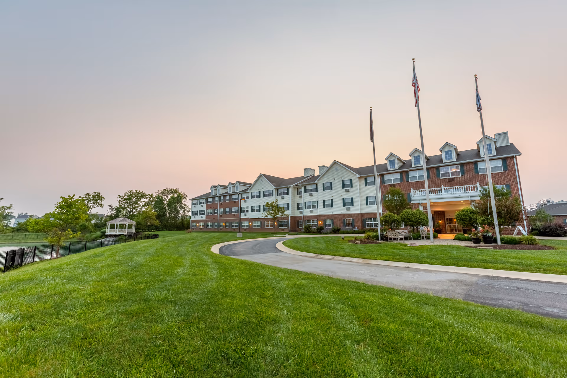 Three-story senior living building with flags, a curved driveway, manicured lawn, and a gazebo by a pond at dusk.