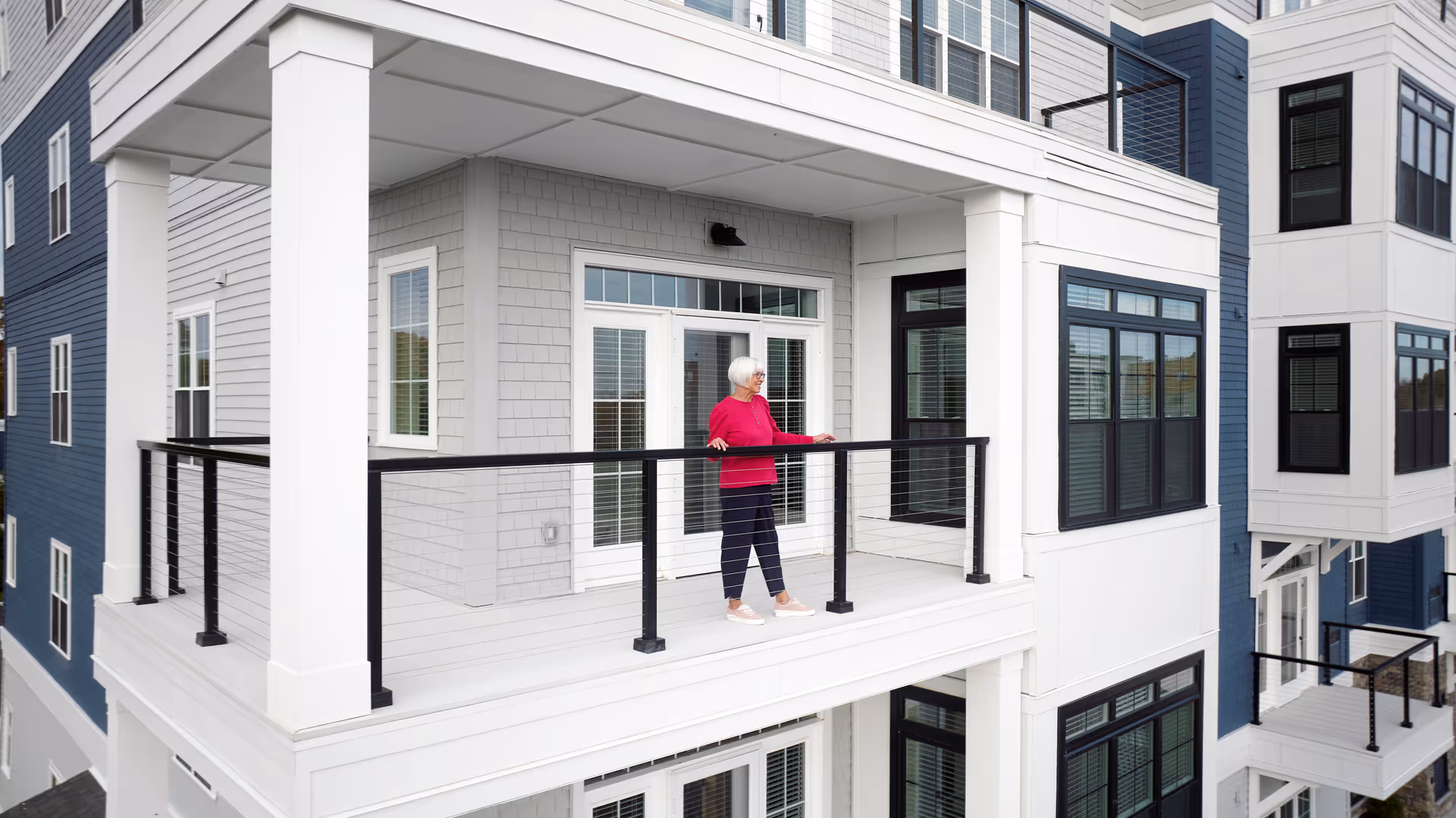 An elderly woman with white hair wearing a red sweater and dark pants stands on a balcony of a modern multi-story building with gray and blue siding and black railings.