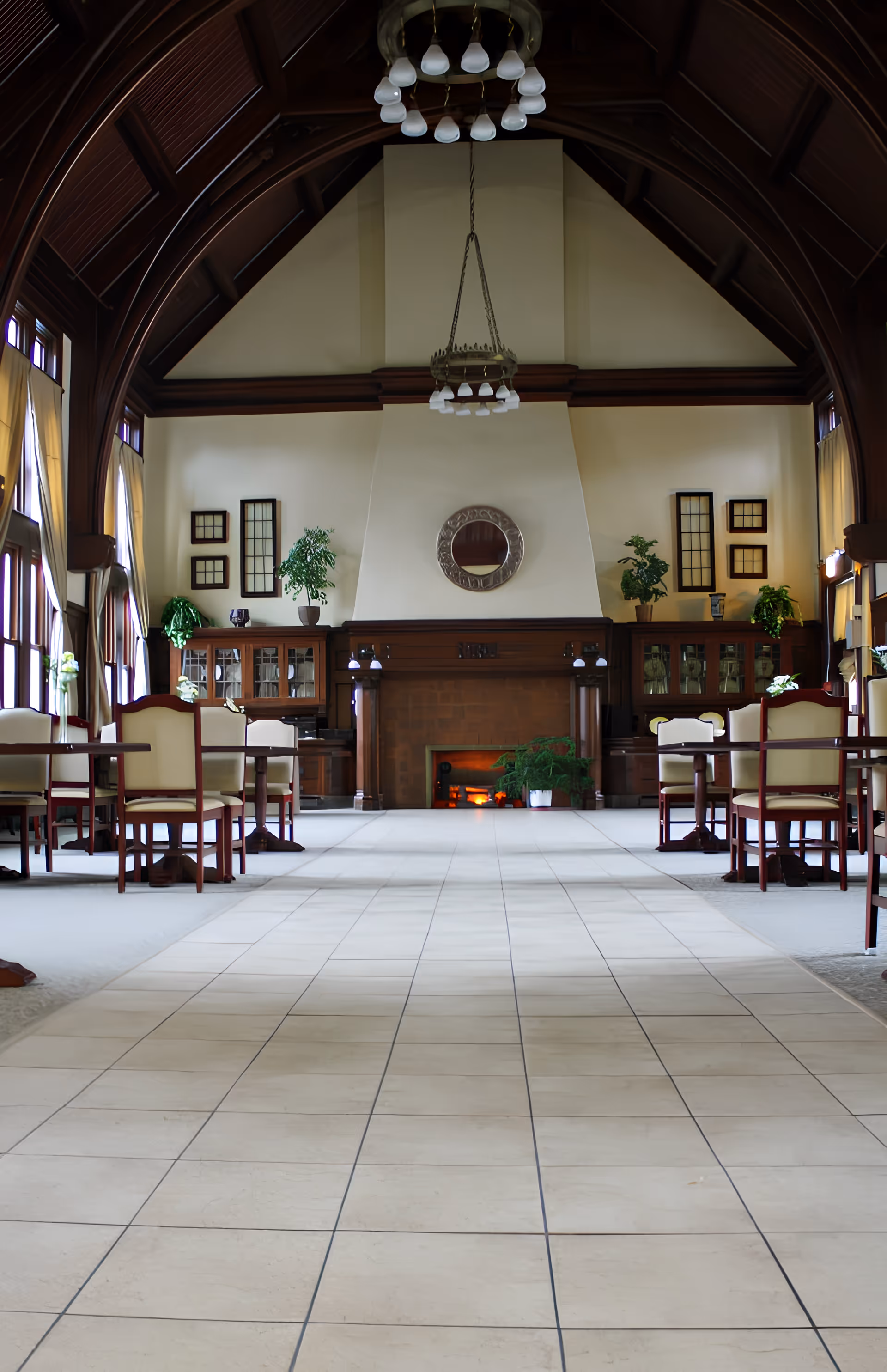 Spacious dining room with a high vaulted wooden ceiling, tiled central aisle, tables and chairs, and a fireplace at the far end.