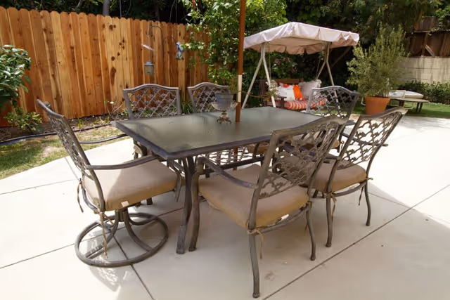 Outdoor patio area with a rectangular glass-top table surrounded by six metal chairs with beige cushions. In the background, there is a cushioned swing with a canopy and decorative pillows, a wooden fence, and some potted plants and greenery.