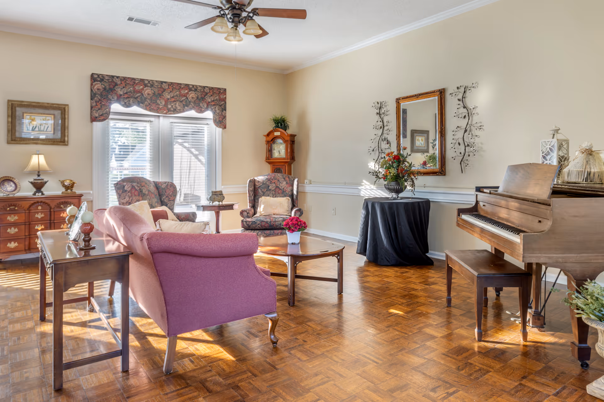 A bright living room with wooden parquet flooring, featuring a pink upholstered sofa, two floral armchairs, a wooden coffee table with a small flower pot, a wooden piano with a bench, a round table covered with a black cloth and a flower arrangement, a grandfather clock, and a ceiling fan with lights. The room has large windows with blinds and a floral valance, and walls decorated with a mirror and metal wall art.