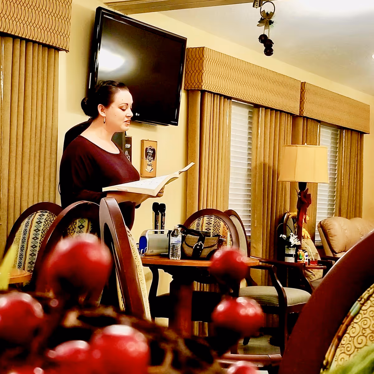 A woman with dark hair tied back is standing in a living room or common area, reading from an open book. The room has patterned chairs, a table with a water bottle and a handbag, a lamp, and windows with beige curtains and valances. A flat-screen TV is mounted on the wall behind her.