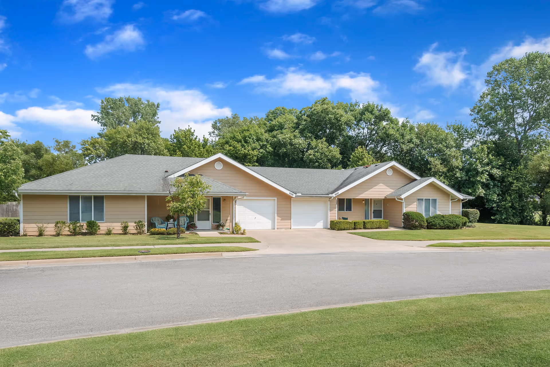 Exterior view of a single-story residential building with beige siding and a gray roof, featuring two attached garages and small front porches with chairs. The building is surrounded by well-maintained green lawns and trees under a blue sky with scattered clouds.