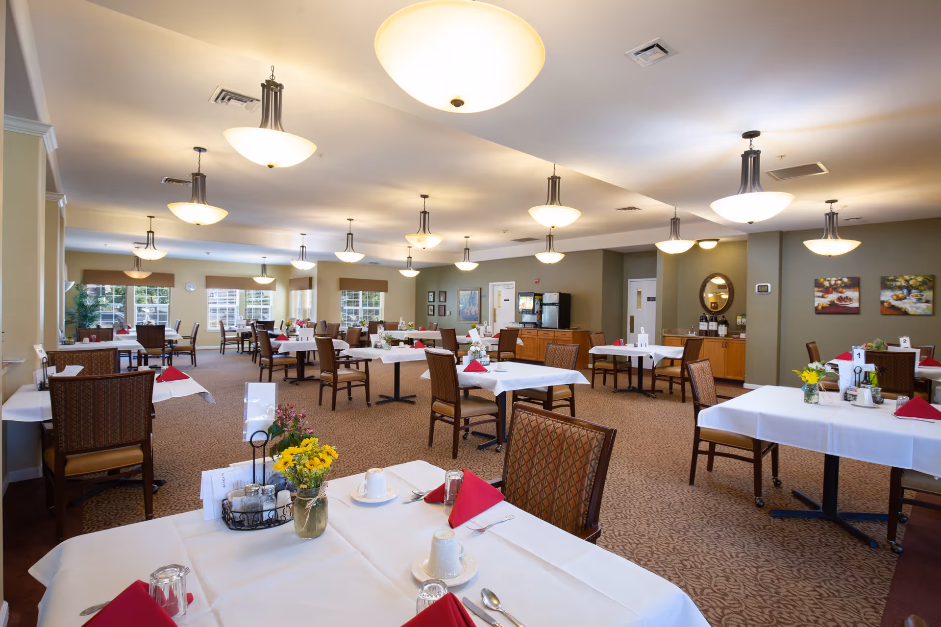 A spacious dining room in a senior living community with multiple tables covered in white tablecloths, each set with red napkins, cups, and silverware. The room is well-lit with ceiling lights and has large windows allowing natural light. There are decorative flowers on the tables and framed artwork on the walls.