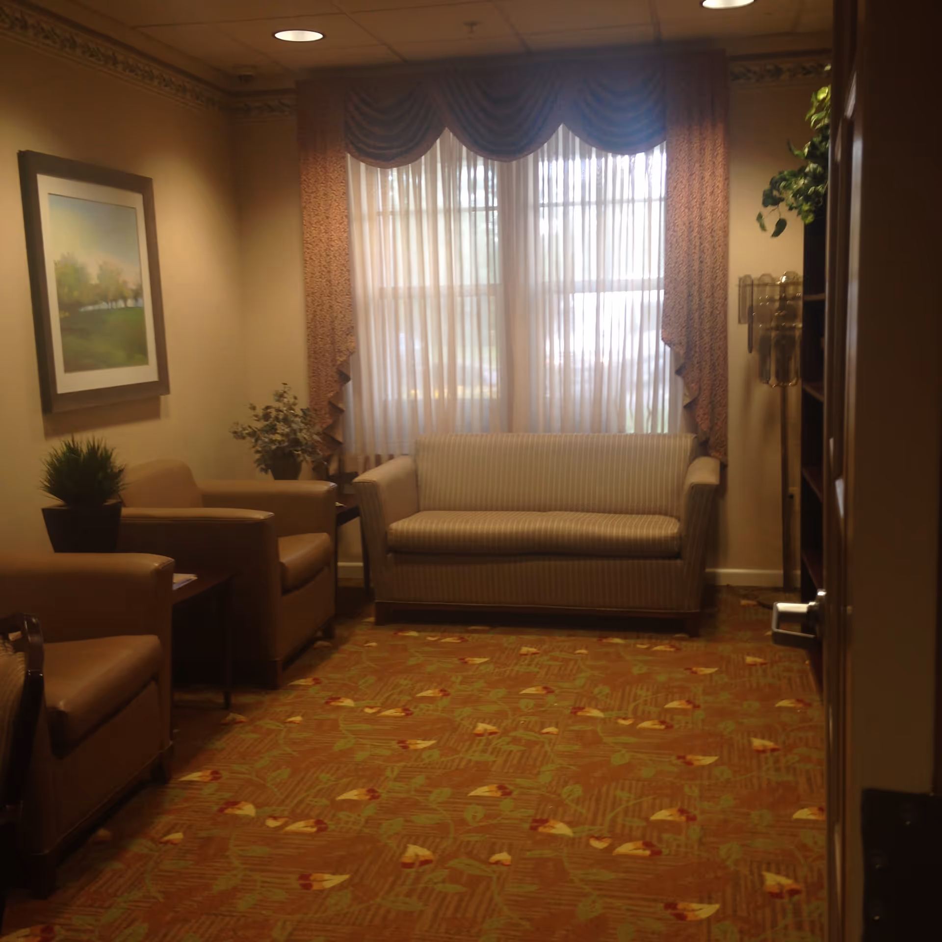 A cozy sitting area with a striped sofa centered under a window with sheer curtains and decorative valance. Two beige armchairs flank a small wooden side table with a potted plant and a floral arrangement. A framed landscape painting hangs on the left wall, and a tall floor lamp stands near a bookshelf on the right. The floor is covered with a patterned carpet featuring floral designs.