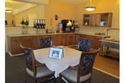 Small dining area with a square table set for four, cushioned chairs, and a serving counter with beverage station and cabinets in the background.