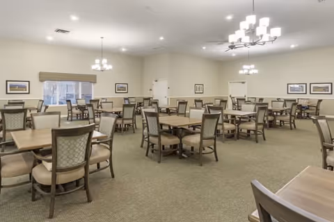 Spacious dining room with multiple wooden tables and chairs, chandeliers, and neutral decor.