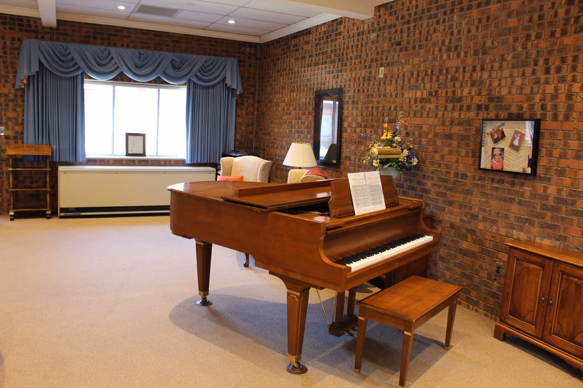 A cozy room with a wooden grand piano and matching bench in the center. The room has brick walls, a beige carpet, and a large window with blue curtains and valance. There is a cushioned armchair with a red pillow, a floor lamp, a wooden cabinet, a framed collage on the wall, and a vase with flowers on a small table.