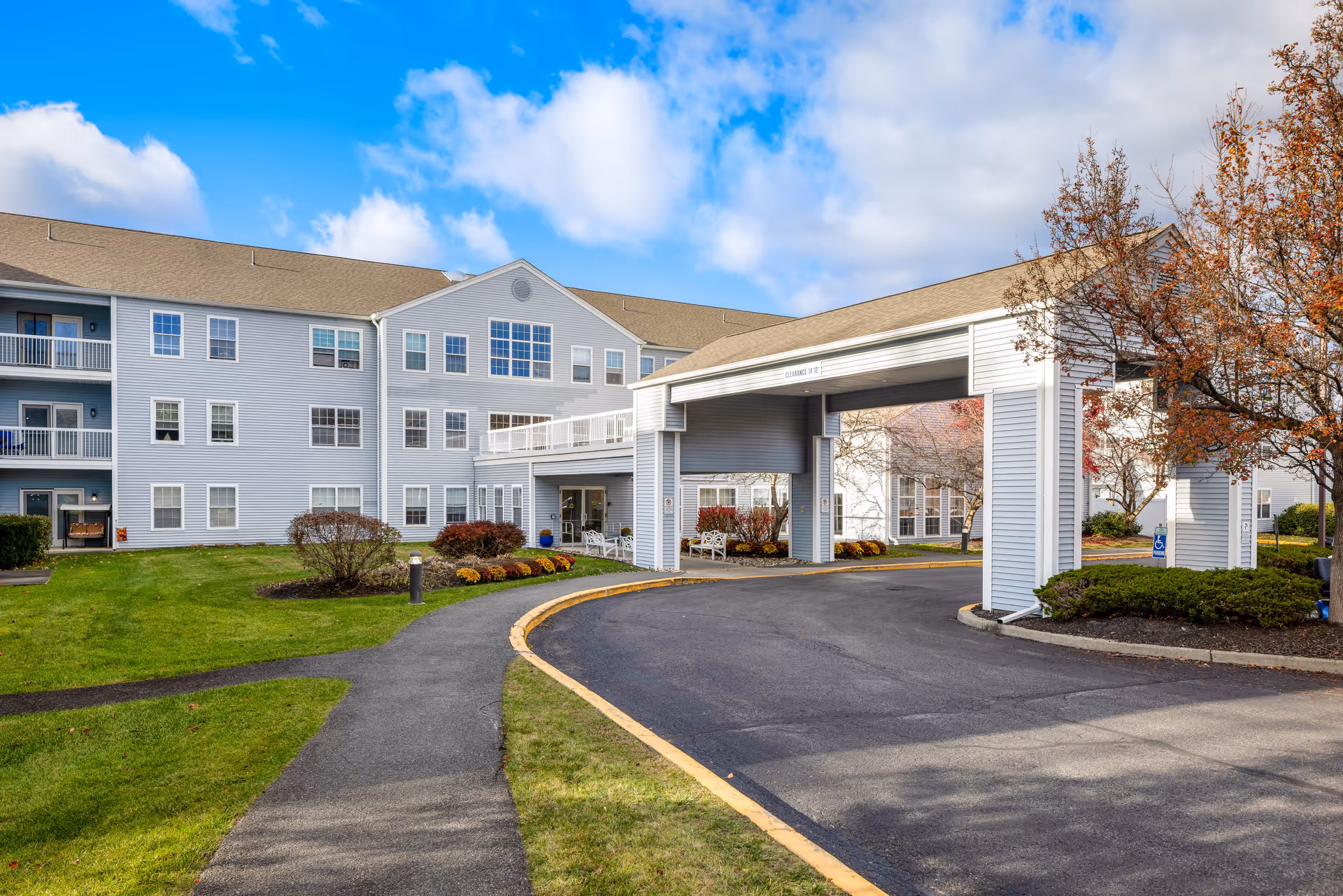 Exterior view of Diamond Ridge - A Provincial Senior Living Community, showing a three-story building with multiple windows and balconies. There is a covered driveway entrance with white pillars and a curved asphalt road leading up to it. The surrounding area includes green lawns, landscaped bushes, and trees with autumn foliage under a partly cloudy blue sky.
