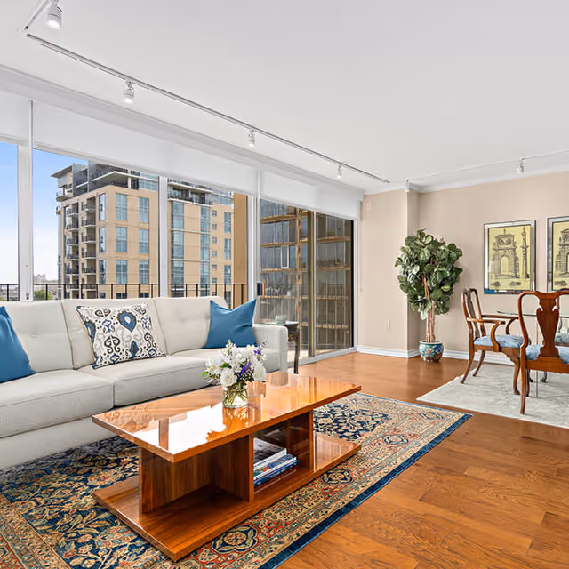 Bright and spacious living room with large floor-to-ceiling windows showing city buildings outside. The room features a white sofa with blue and patterned pillows, a wooden coffee table with a flower vase, a patterned area rug, a dining table with chairs, two framed artworks on the wall, and a tall potted plant in the corner.