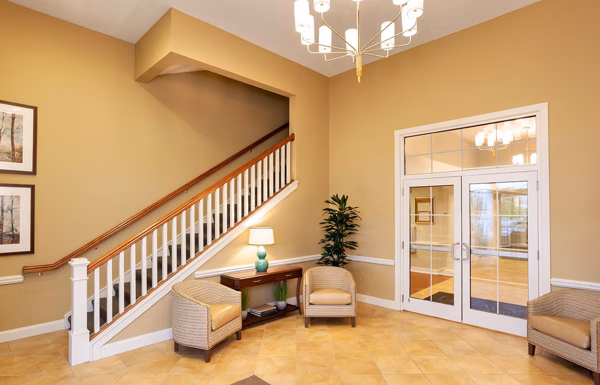 A cozy interior seating area with two cushioned armchairs and a small wooden table with a lamp and decorative plants. There is a staircase with a wooden handrail and white balusters on the left side, and double glass doors with white frames on the right. The walls are painted beige, and the floor is tiled in a light tan color. Two framed pictures hang on the wall near the staircase, and a modern chandelier hangs from the ceiling.