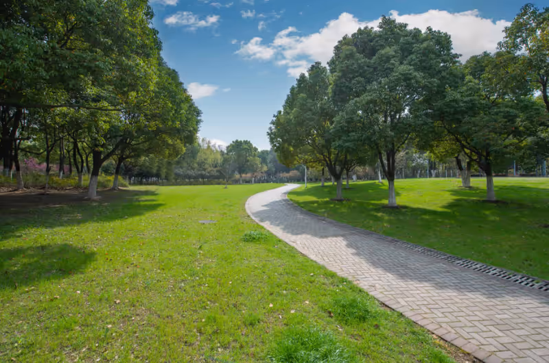 A paved winding pathway through a green park area with well-maintained grass and numerous leafy trees under a partly cloudy blue sky.