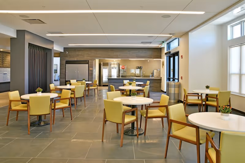 A bright and modern dining area with round white tables and yellow cushioned chairs. Small potted plants are placed on each table. The back of the room features a serving counter with kitchen appliances and a refrigerator. Large windows on the right side allow natural light to fill the space.