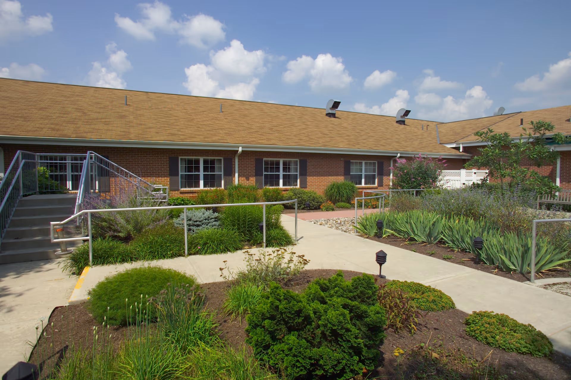 Outdoor courtyard area of a nursing home with a paved walkway, handrails, stairs, and various green plants and shrubs. The building has a brick exterior with several windows and a tan shingled roof under a partly cloudy sky.