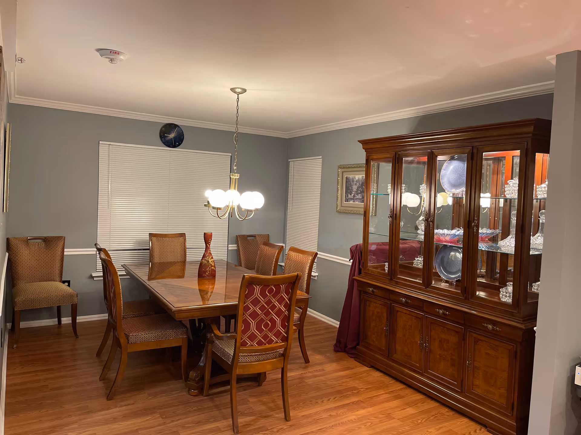 A dining room with a wooden dining table surrounded by eight chairs, a chandelier hanging above the table, a wooden china cabinet displaying glassware and plates, and two windows with closed blinds. The walls are painted gray and the floor is hardwood.