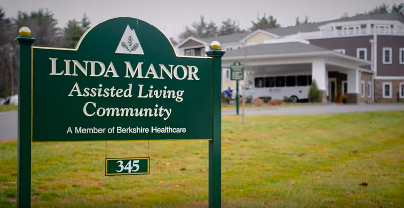 Green sign reading "Linda Manor Assisted Living Community" with the facility building and covered entrance visible in the background.