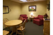 A small living room area with two red upholstered armchairs, a wooden side table with a lamp, a round wooden dining table with four chairs, a framed picture on the wall, and a wooden cabinet with a plant on top.