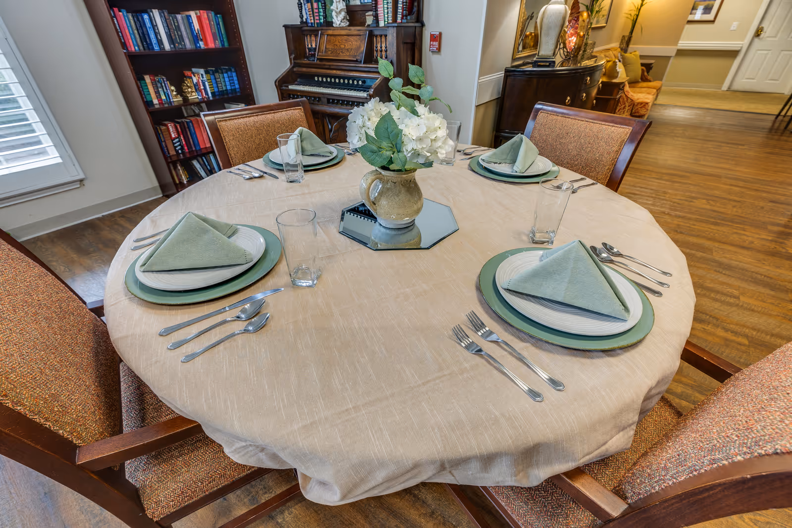 A round dining table set for four with beige tablecloth, green plates, white napkins folded in a triangle, clear drinking glasses, and silverware. A vase with white flowers is placed in the center on a mirrored base. The room has wooden flooring, a bookshelf filled with books, and an antique organ in the background.