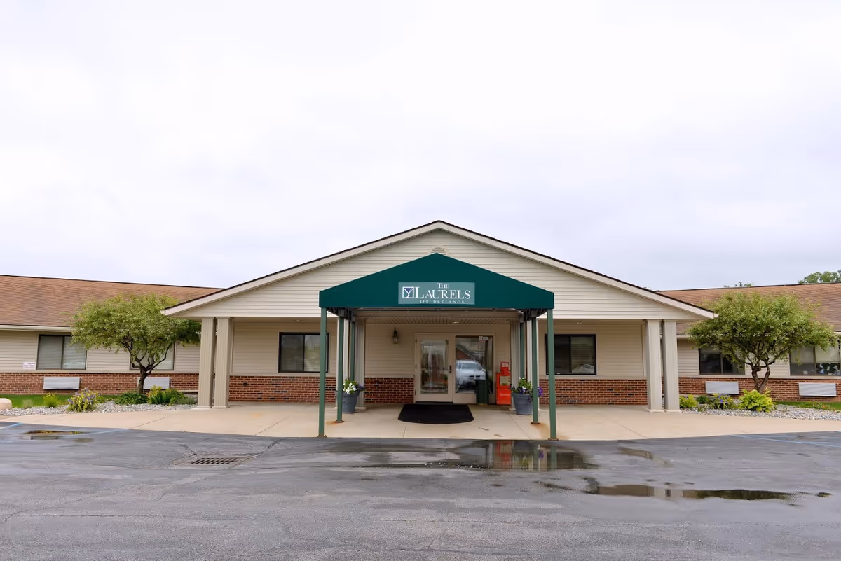 Front exterior view of The Laurels of Defiance facility with a green awning over the entrance, two small trees on either side, and a paved driveway with some puddles in front.