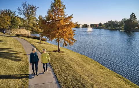 Two elderly women walking and talking on a paved path beside a lake with a fountain, surrounded by green grass and trees under a clear blue sky.
