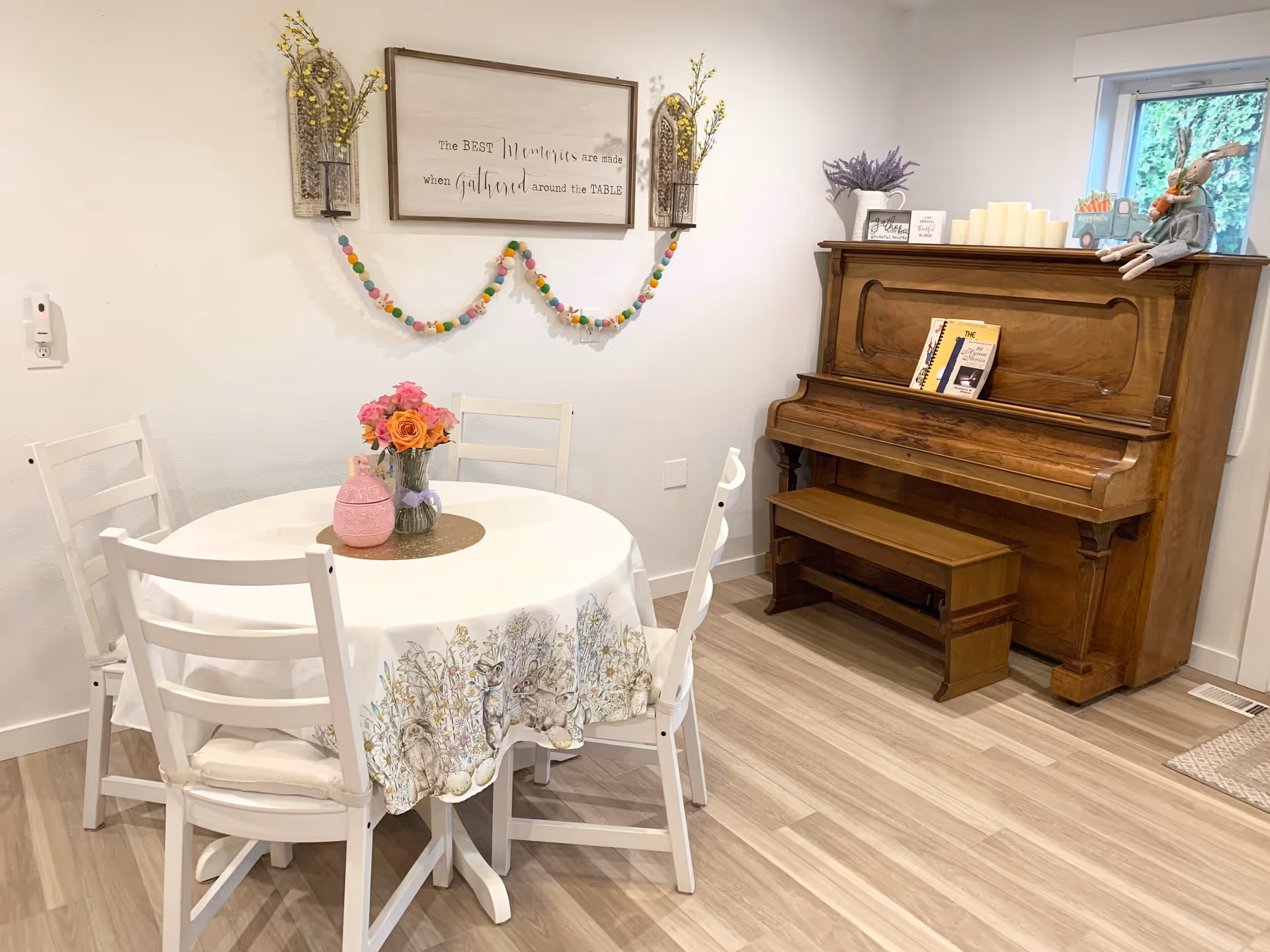 A cozy interior room featuring a round table covered with a white tablecloth decorated with floral and bunny designs. Four white chairs surround the table. On the table, there is a vase with colorful flowers and a pink decorative jar. Against the wall is a wooden upright piano with a matching bench, topped with candles, a small sign, and a stuffed bunny doll. Above the table, a framed sign reads 'The BEST Moments are made when gathered around the TABLE' with decorative wall hangings and a colorful garland. A small window is visible near the piano, letting in natural light.