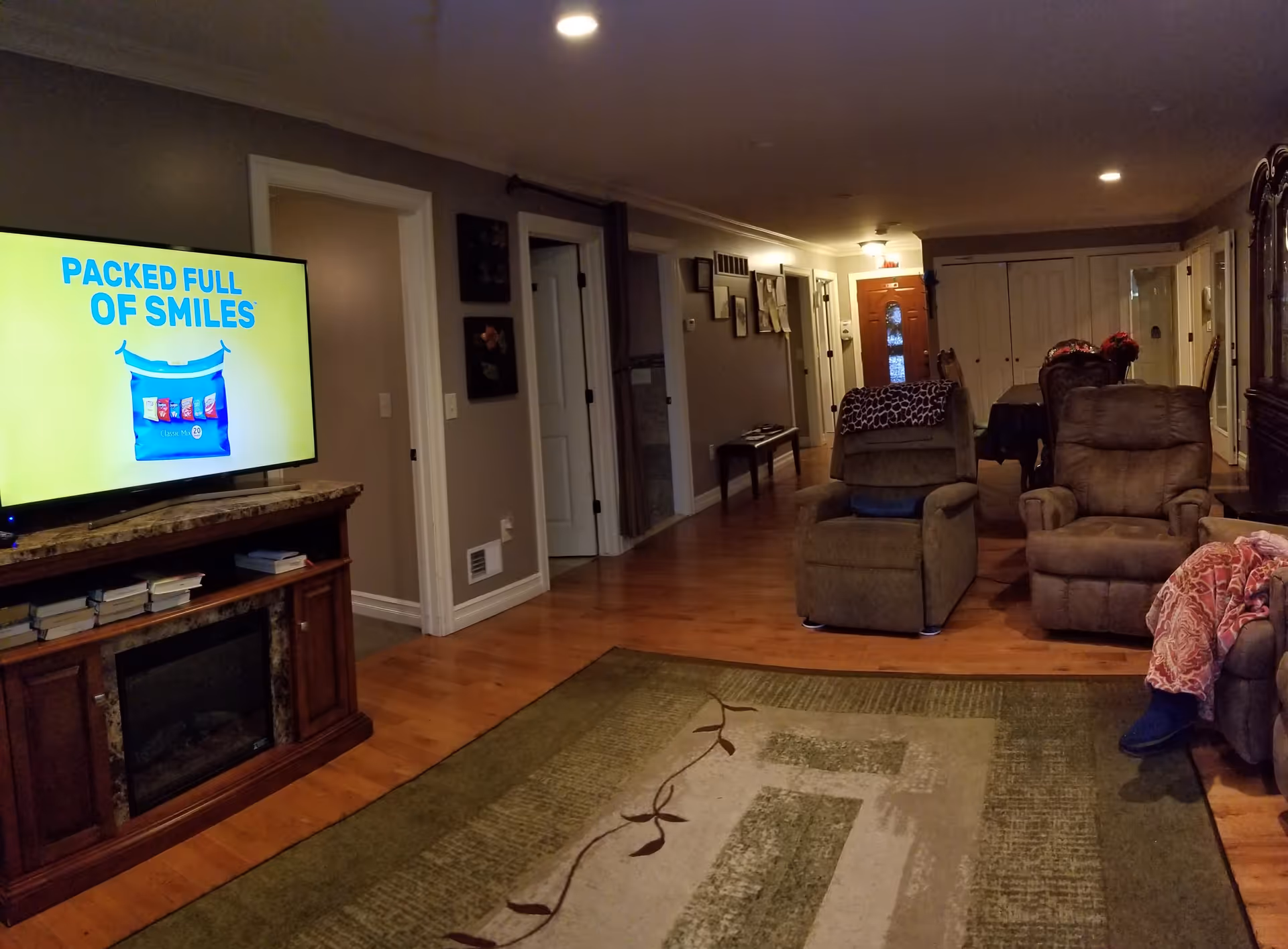 A cozy living room with two recliner chairs, a large area rug, a wooden TV stand with a flat-screen TV displaying an advertisement that reads 'PACKED FULL OF SMILES'. The room has wooden flooring, a hallway with framed pictures on the wall, and a dining table with chairs in the background.