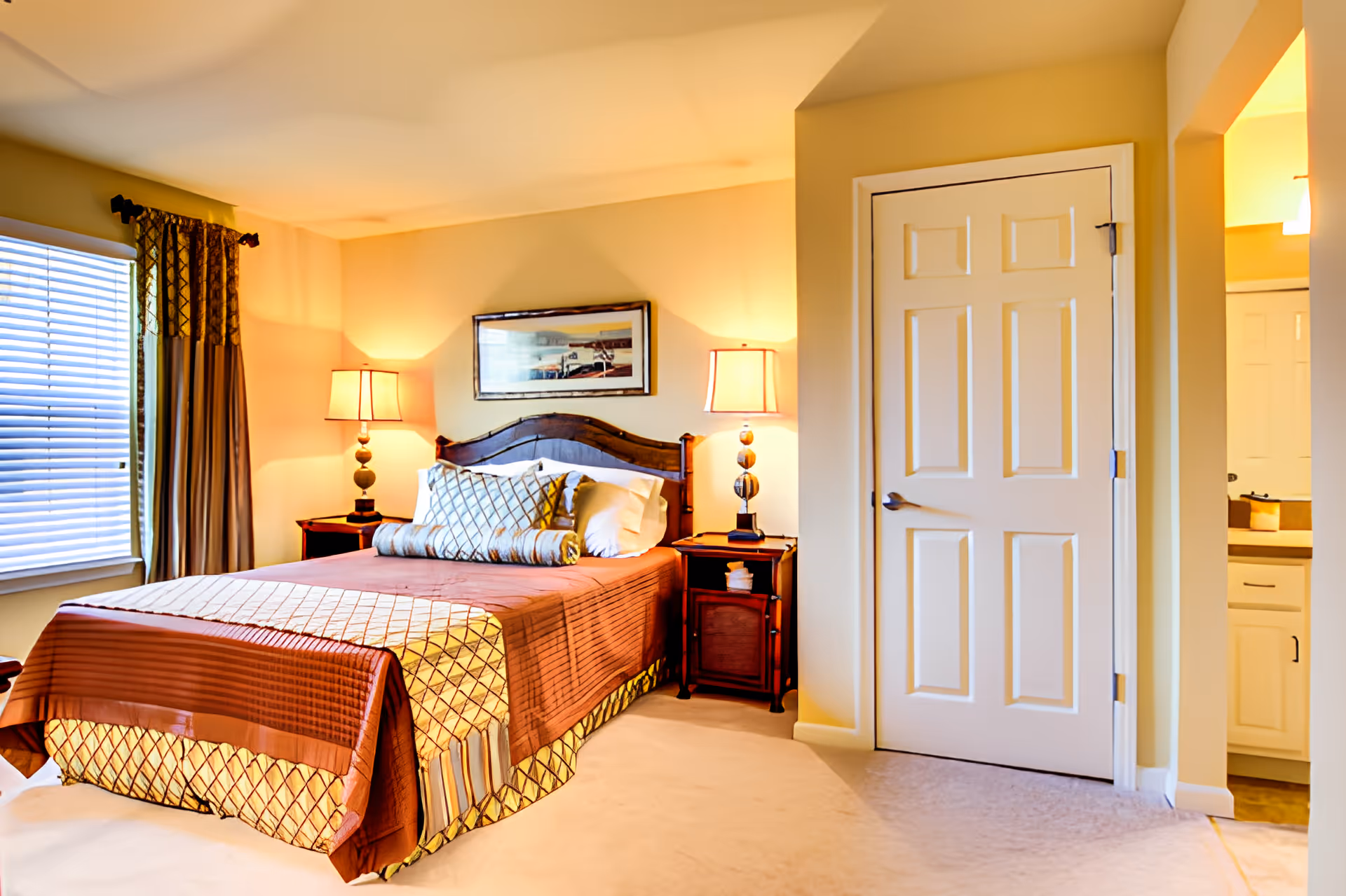 A warmly lit bedroom with a neatly made bed featuring brown and beige bedding, two bedside tables each with a lamp, a window with curtains on the left, and a closed white door on the right. A framed picture hangs above the bed.