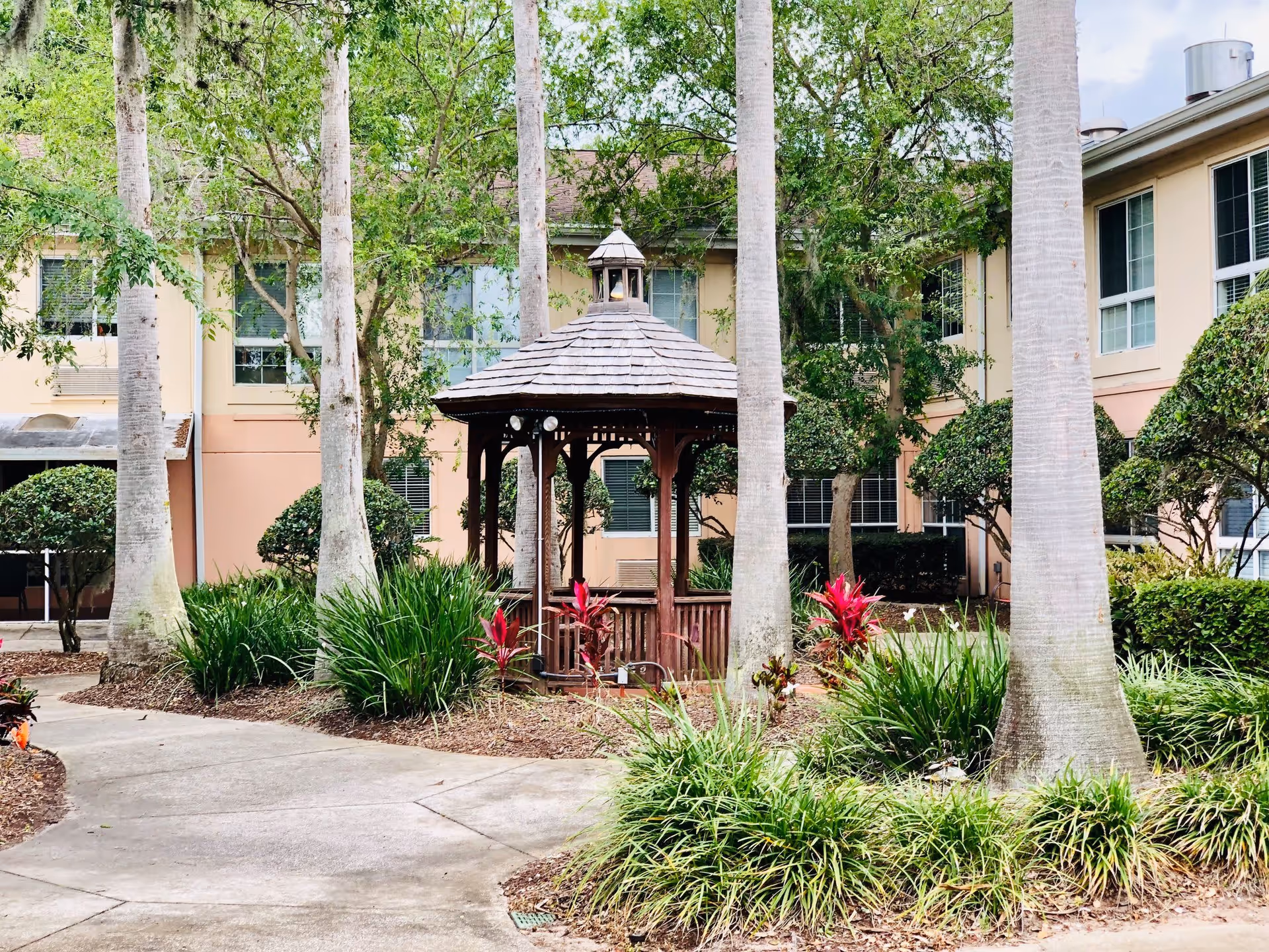 Outdoor courtyard area at Ansley Cove featuring a wooden gazebo surrounded by palm trees, green shrubs, and red flowering plants, with a beige multi-story building in the background.