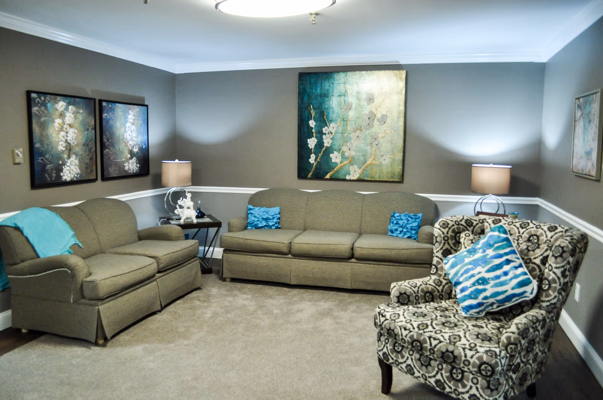 A cozy living room with two beige sofas and a patterned armchair with a blue and white pillow. The walls are painted gray with white trim, and there are three floral paintings hanging on the walls. Two table lamps with beige shades are placed on side tables beside the sofas, providing soft lighting.
