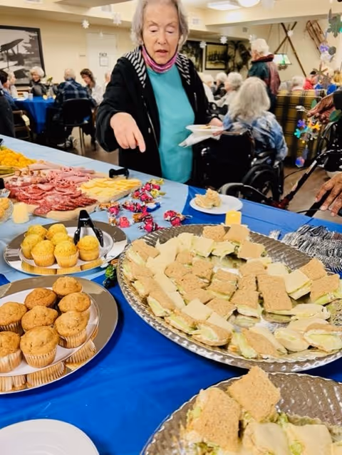 An elderly woman points at a buffet table of sandwiches and muffins in a dining room with other residents seated in the background.