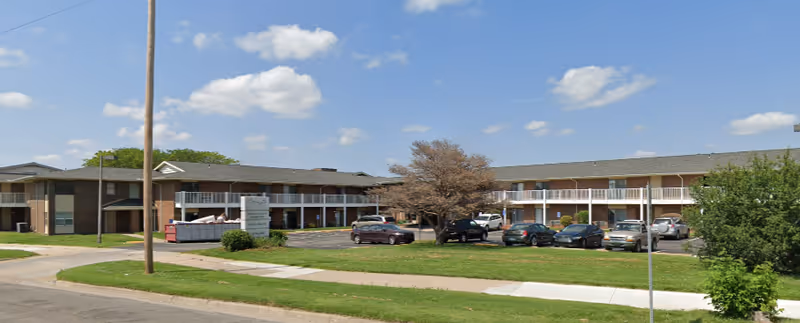 Exterior view of a two-story senior living facility with a parking lot in front, several parked cars, a tree with sparse leaves, and a sign near the entrance. The building has balconies on the second floor and a clear blue sky with some clouds above.