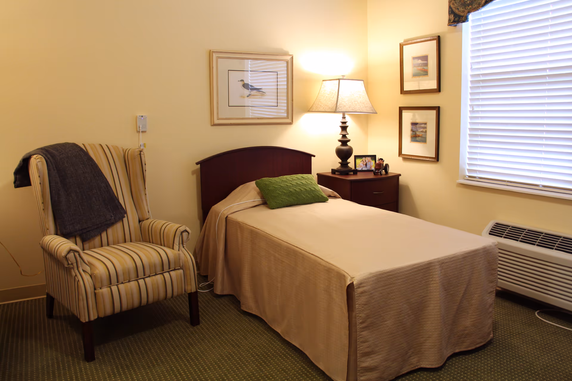 A cozy bedroom with a single bed covered in a beige bedspread and a green pillow. Next to the bed is a wooden nightstand with a lamp, a framed photo, and some small items. A striped armchair with a dark throw blanket draped over the back is positioned to the left of the bed. The walls are light yellow, decorated with framed pictures, and there is a window with closed blinds on the right side. A heating or air conditioning unit is below the window.