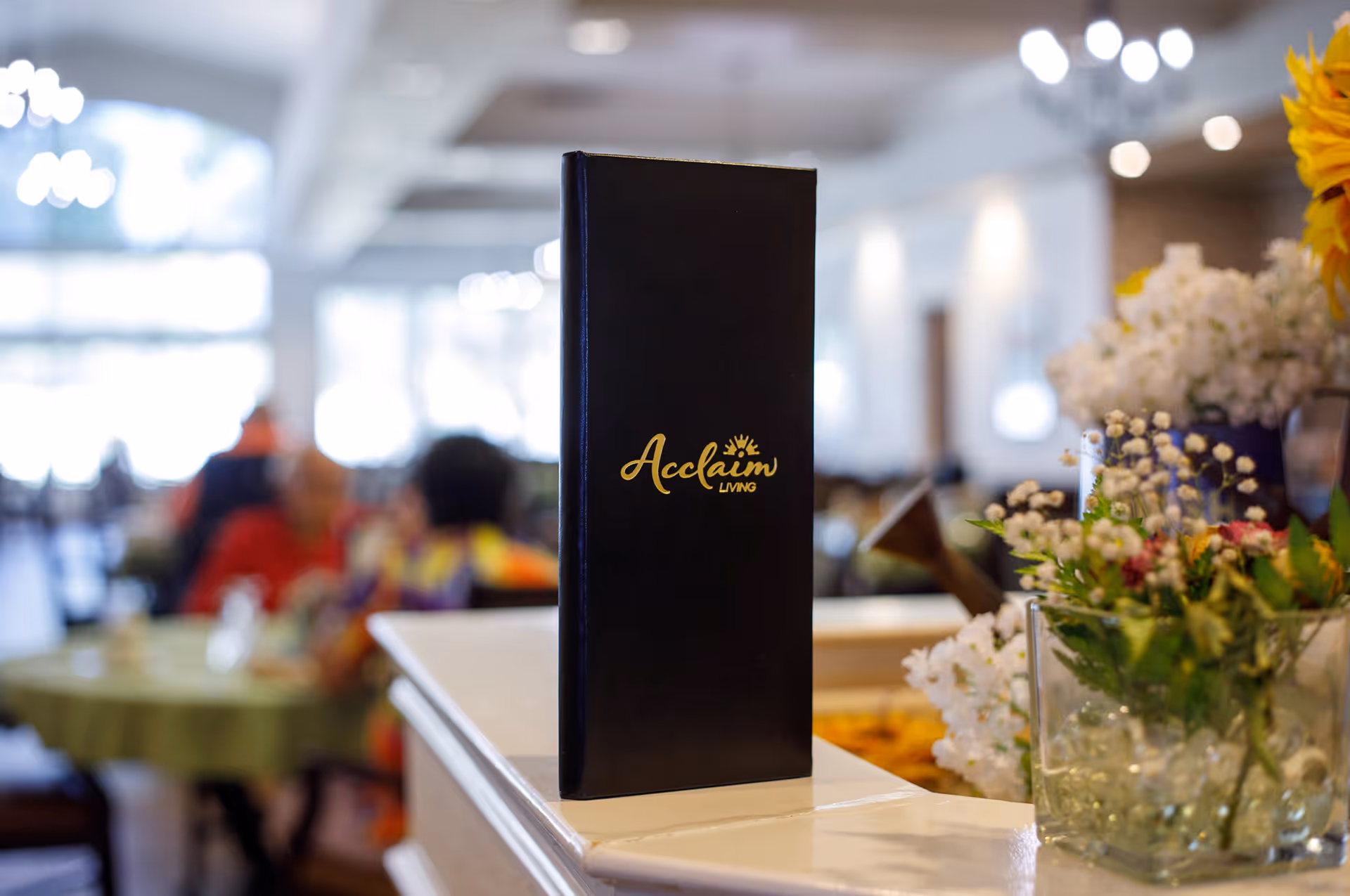 A black menu or brochure with the gold text 'Acclaim Living' standing upright on a white counter. In the background, there is a softly focused dining area with people sitting at tables and floral arrangements on the counter.