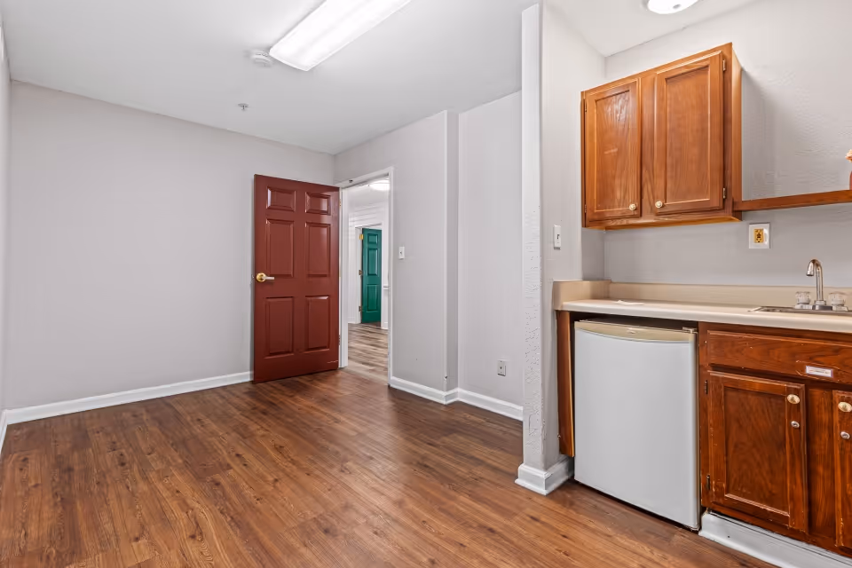 An empty room with wood flooring and light gray walls featuring a small kitchenette with wooden cabinets, a countertop, a sink, and a mini refrigerator. A red door is open, revealing a hallway with a green door in the background. The room is lit by a fluorescent ceiling light.