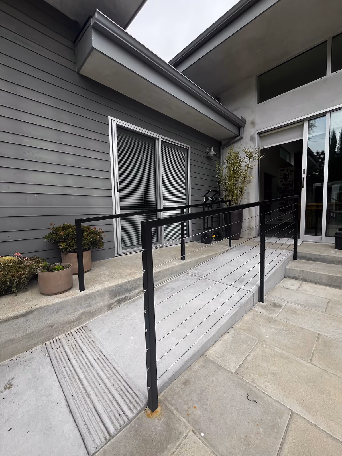 Concrete wheelchair ramp with black cable railing leading to a modern home's sliding glass door and potted plants.