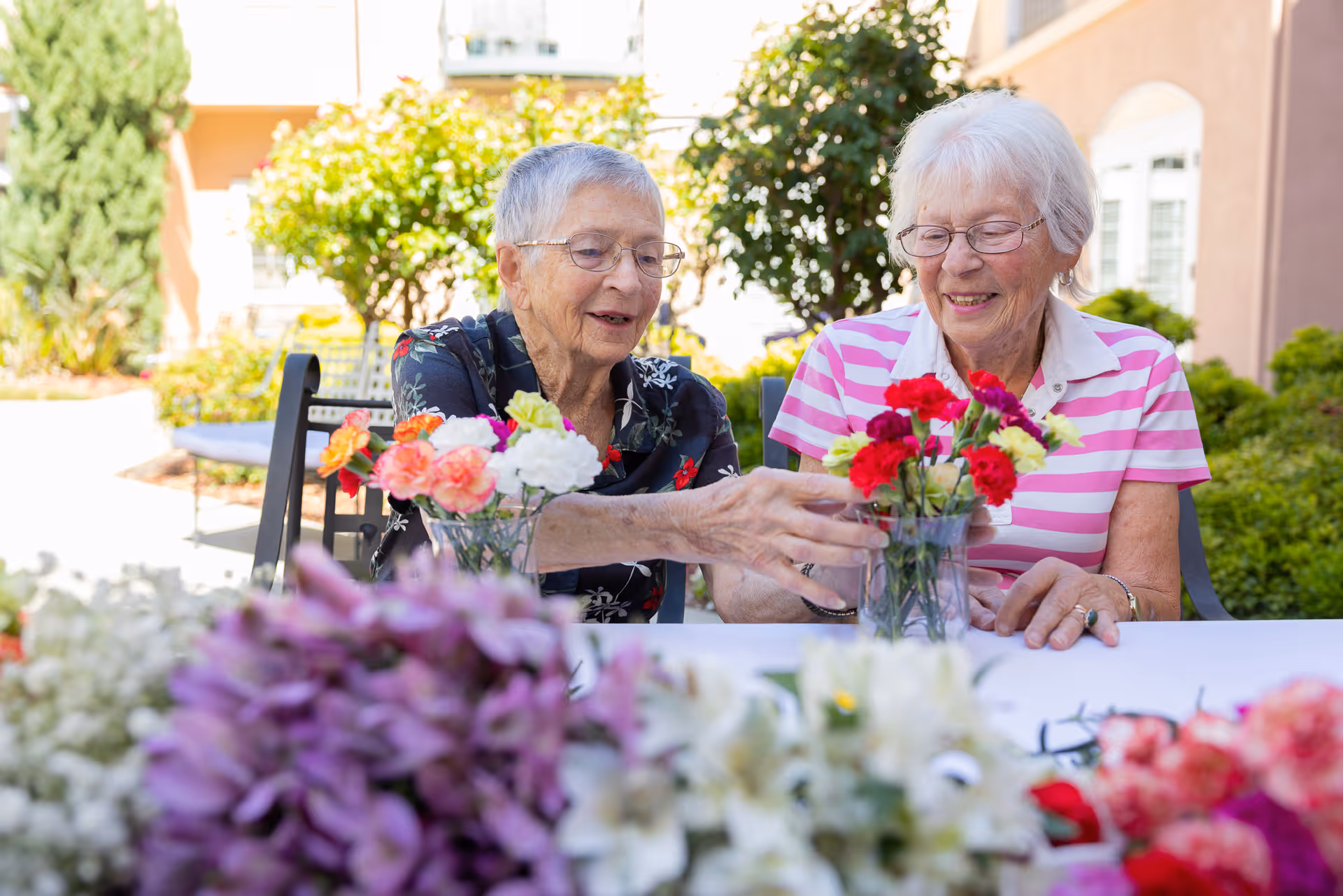 Two elderly women sitting at a table outdoors arranging colorful flowers in glass vases, surrounded by greenery and buildings in the background.