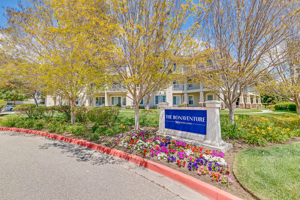 Exterior view of a senior living facility named The Bonaventure with a landscaped garden featuring colorful flowers and trees with light green leaves. The building is a multi-story structure with balconies and large windows. A blue sign with white text reads 'THE BONAVENTURE SKY ACTIVE LIVING' in front of the building.