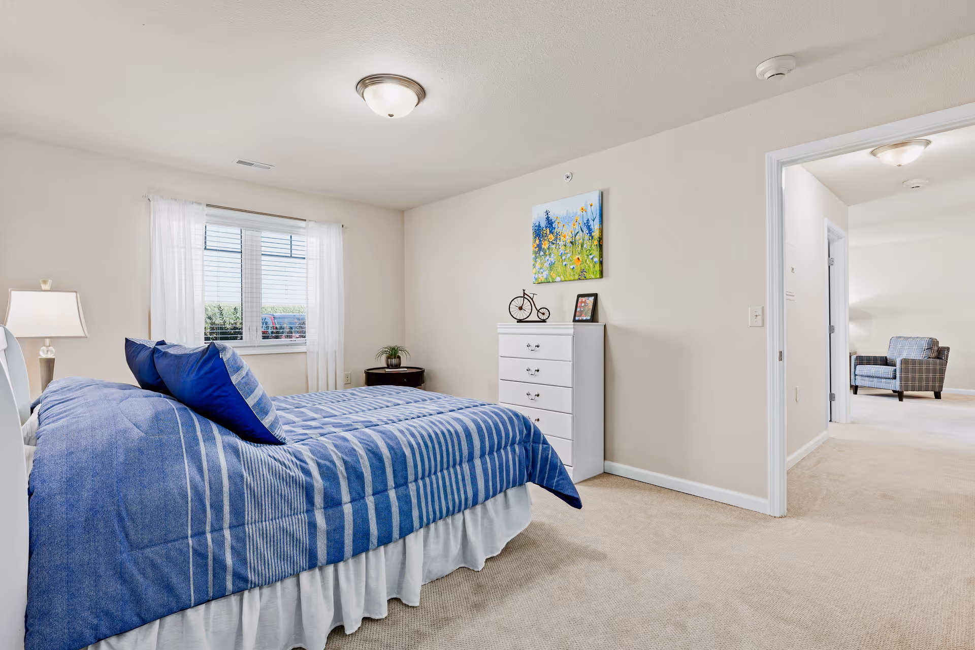 Bright bedroom featuring a blue-striped bed, white dresser, and a window with sheer curtains opening to a hallway and sitting area.