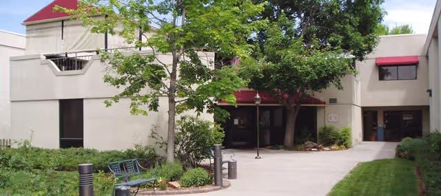 Entrance courtyard of Cherrelyn Health Care Center showing the building facade, walkway, trees, benches, and landscaping.