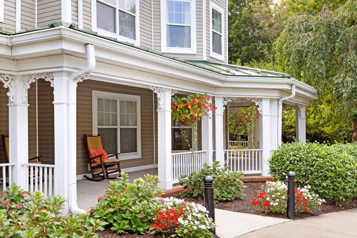Front porch of a senior living facility with white columns, hanging flower baskets, rocking chairs, and well-maintained garden beds with red and white flowers and green shrubs.