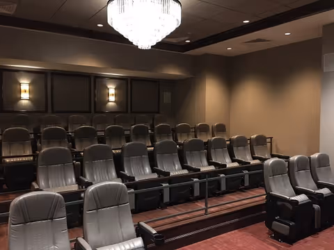 A small theater room with three rows of gray cushioned recliner seats arranged in tiers. The room has dim lighting with wall sconces and a large chandelier hanging from the ceiling. The walls are painted in neutral tones.