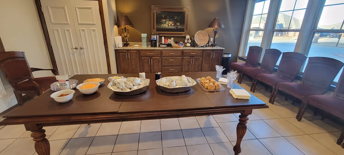 Long wooden table set with covered trays, bowls and muffins in a community dining/refreshment area with chairs along windows and a countertop coffee station.
