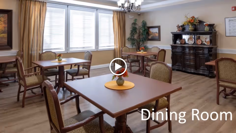 A dining room with several wooden tables and cushioned chairs arranged neatly. Each table has a small vase with flowers on a yellow placemat. The room has large windows with blinds and curtains, a chandelier hanging from the ceiling, and a dark wooden cabinet decorated with plates and floral arrangements. There are framed paintings and a potted plant in the corner.