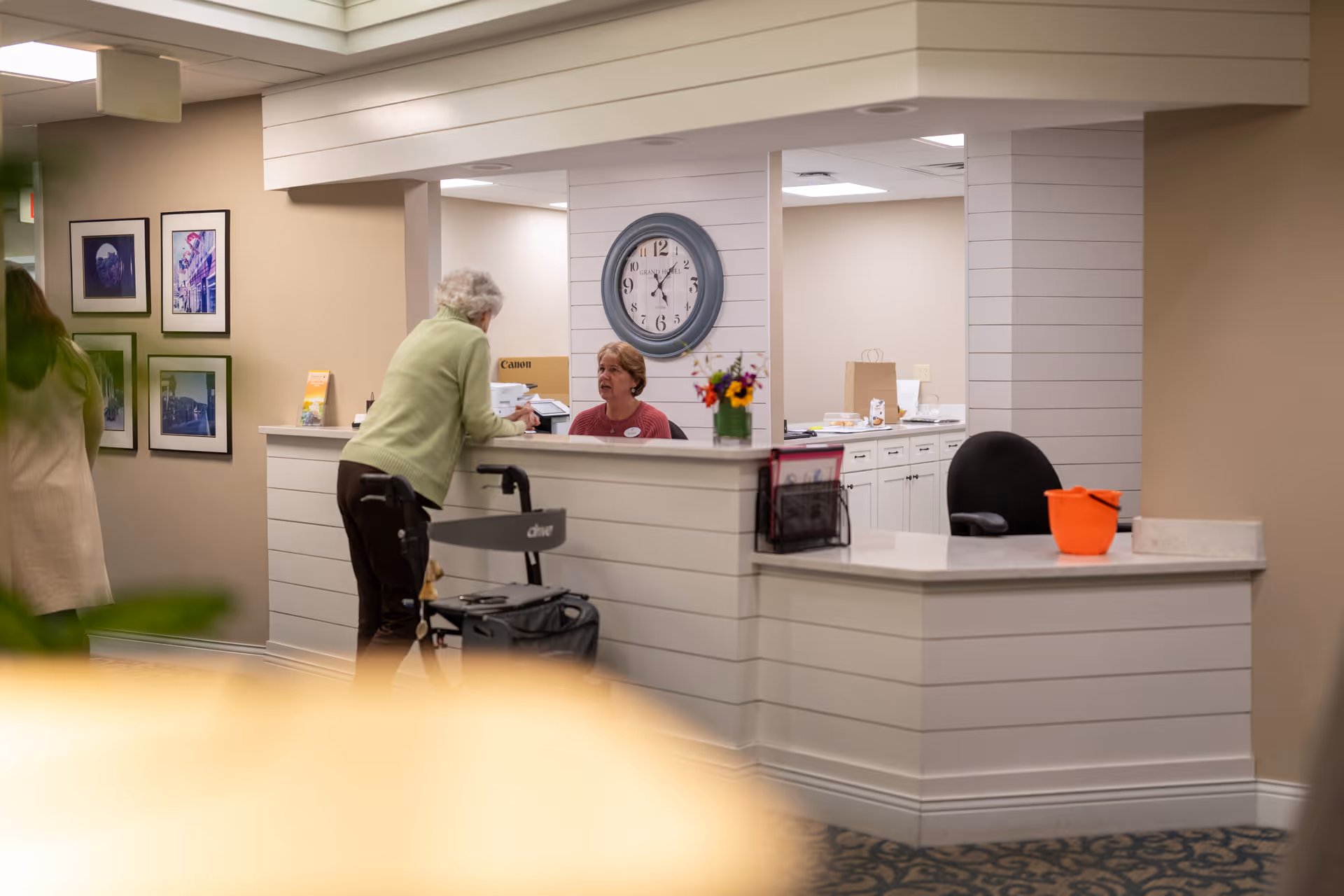 An elderly woman with a walker is talking to a receptionist at a white paneled front desk in a senior living facility. The receptionist is seated behind the desk with a clock on the wall behind her. There are framed pictures on the wall to the left and a small vase with flowers on the desk.