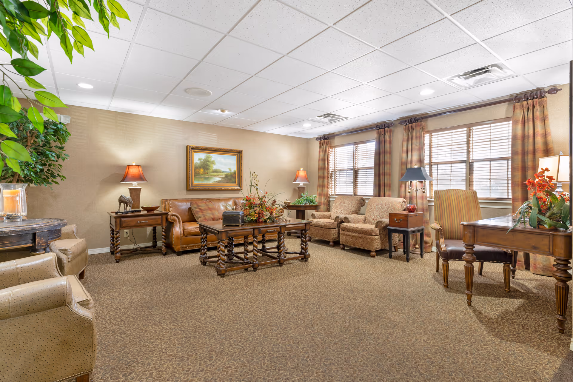 Well-lit common room with sofas, armchairs, coffee tables, lamps, and large windows with curtains.