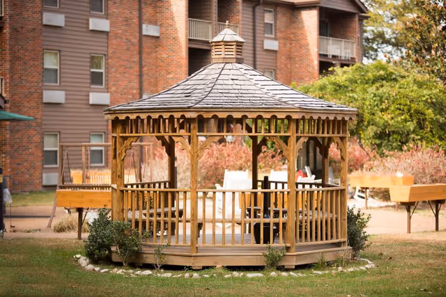 A wooden gazebo with a shingled roof situated on a grassy area in front of a brick residential building. The gazebo is surrounded by small plants and rocks, with outdoor benches and greenery visible in the background.