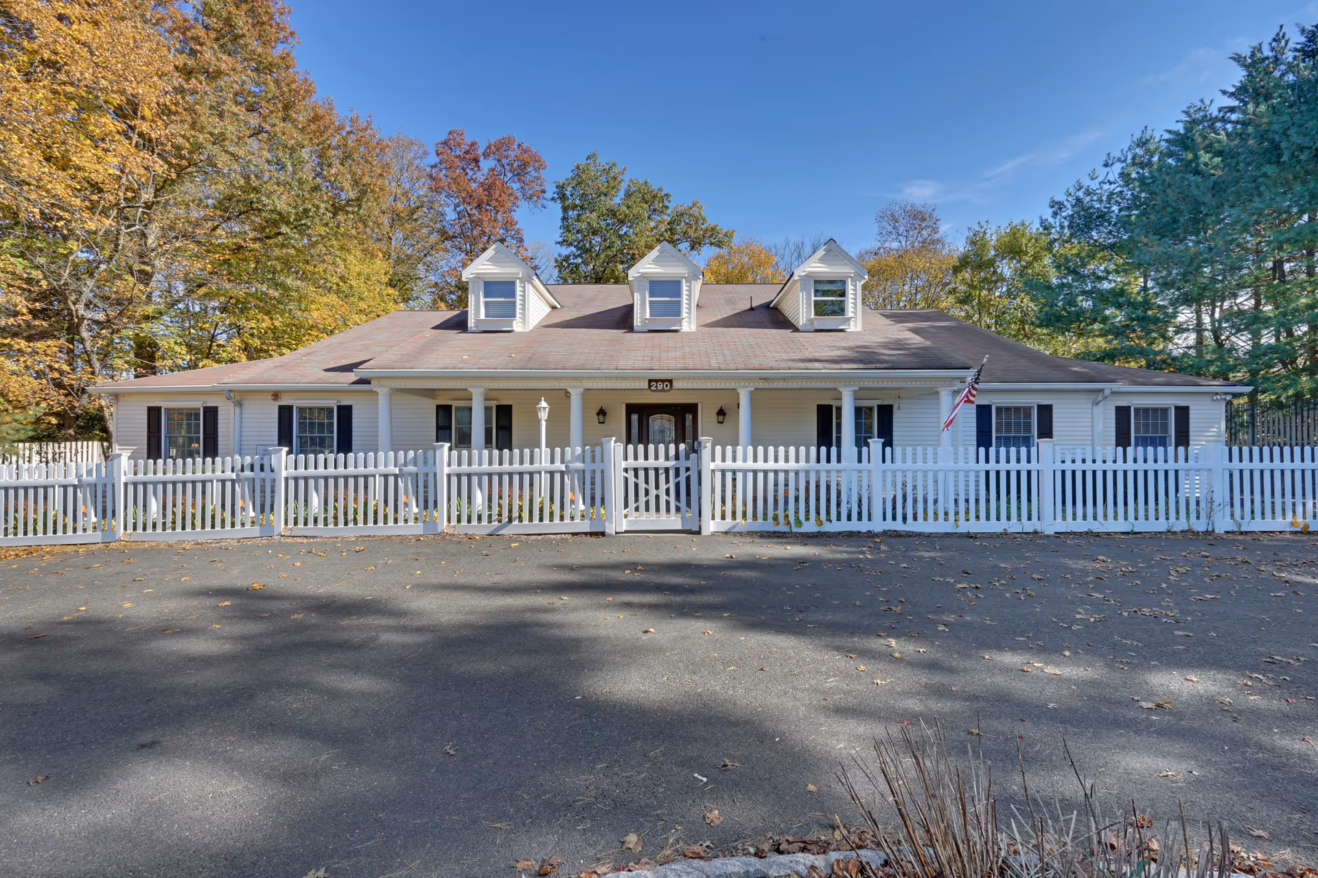 Front exterior view of a single-story building with white siding, three dormer windows on the roof, and a white picket fence enclosing the front yard. There is an American flag mounted near the entrance, and trees with autumn foliage surround the building under a clear blue sky.
