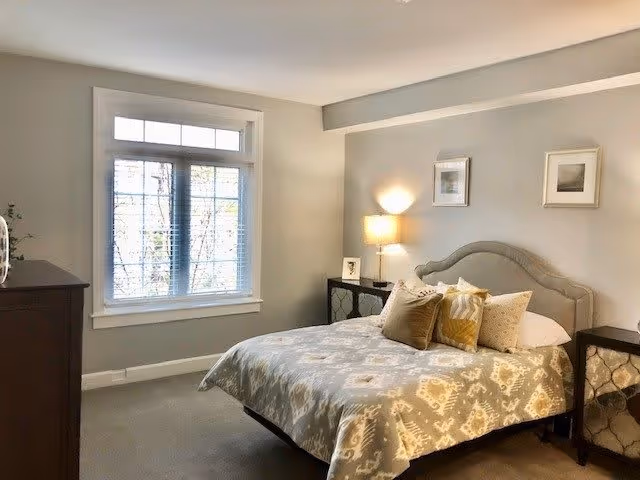 A cozy bedroom with a large window letting in natural light. The room features a bed with a patterned bedspread and multiple pillows, two bedside tables with lamps, and framed pictures on the wall. The walls are painted light gray and the carpet is beige.
