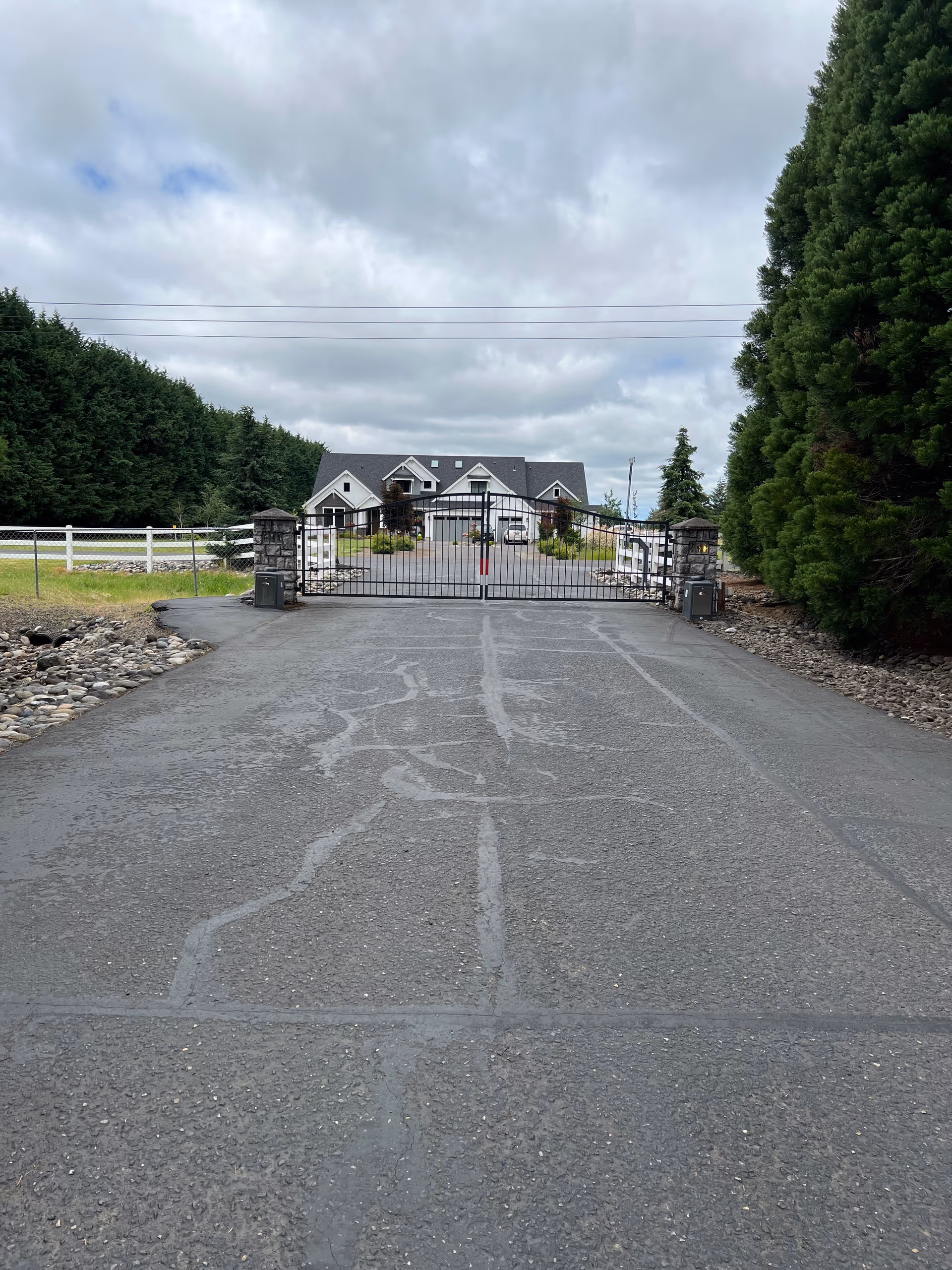 Gated driveway leading to a large house with stone pillars, white fence, and tall trees under a cloudy sky.